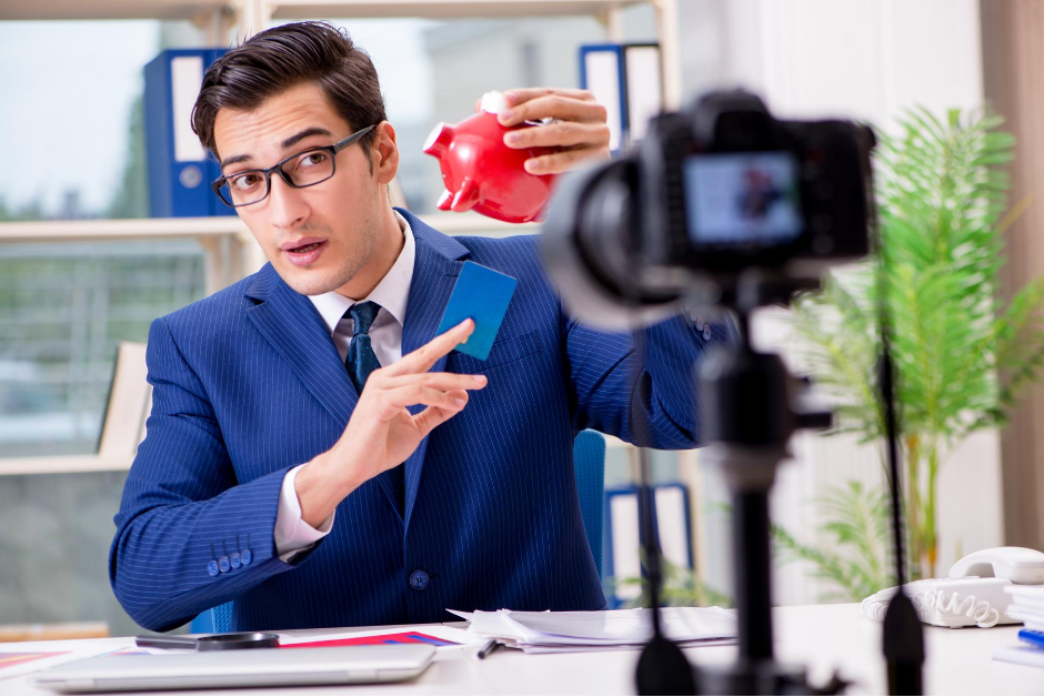 Image of man pointing to a piggy bank who is being filmed by a camera.